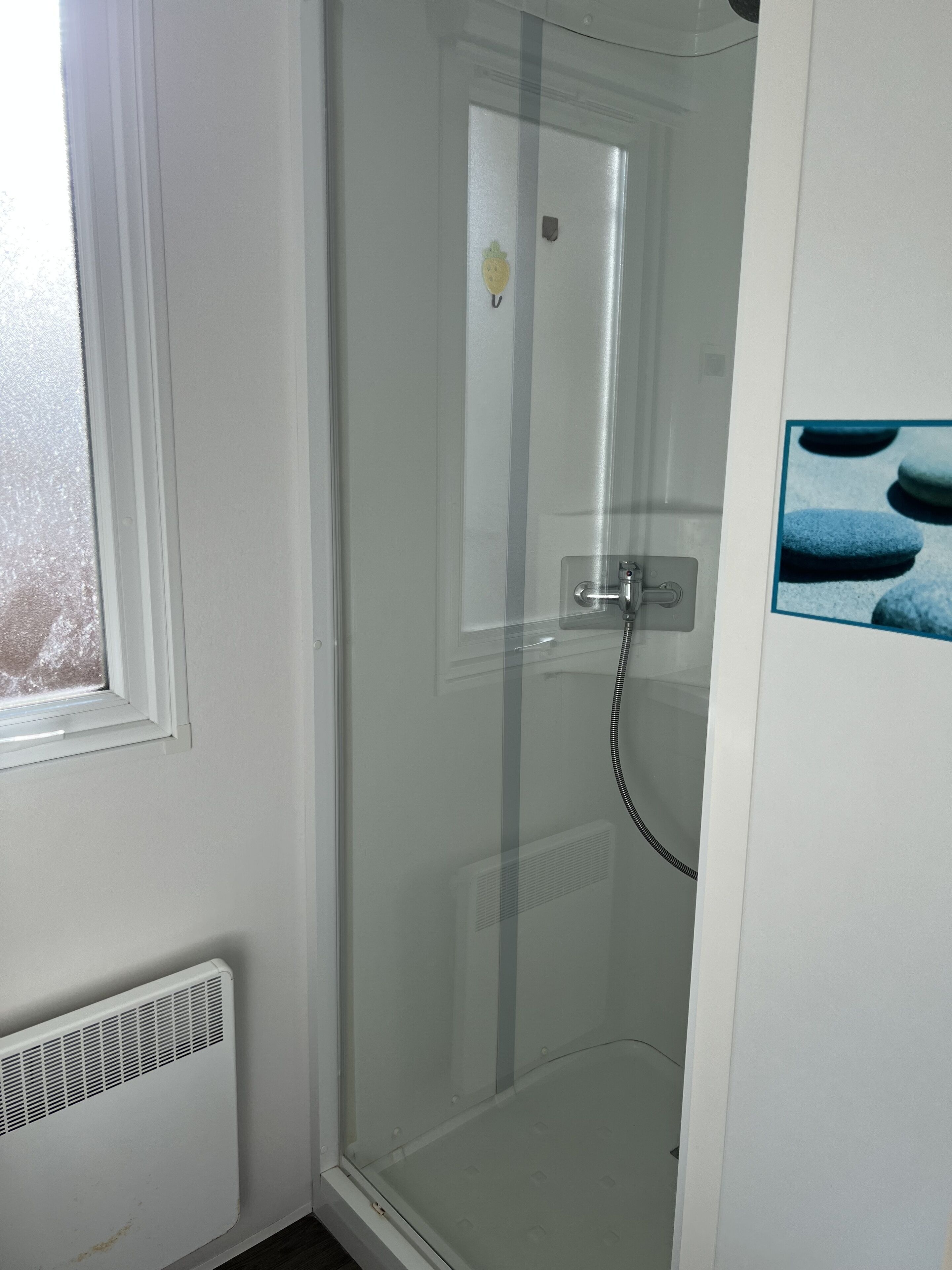 A glass-enclosed shower stall with a white door, a vent below, and a small picture of blue stones on the wall.