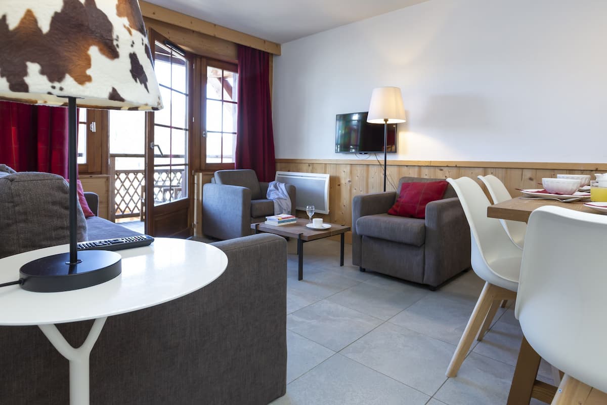 A modern living room with a sofa, armchair, dining table, and a TV mounted on the wall.