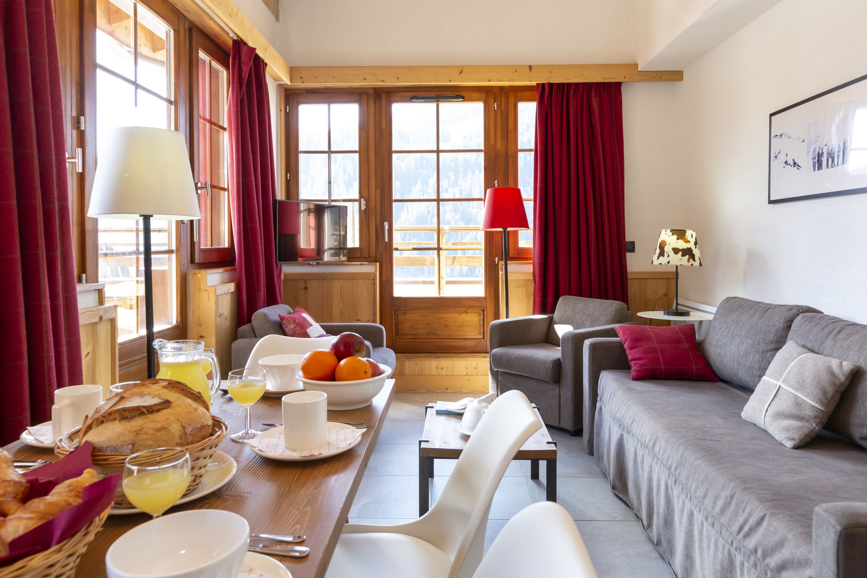 Una sala de estar acogedora con un sofá, una mesa de comedor con alimentos para el desayuno y vistas a las montañas a través de amplios ventanales.