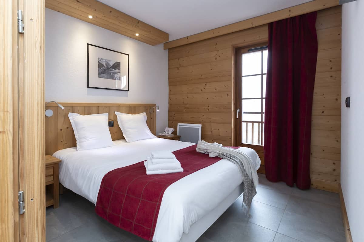 A bedroom with a wooden headboard, a bed with white bedding and a red blanket, a bedside table with a lamp, and a window with red curtains.