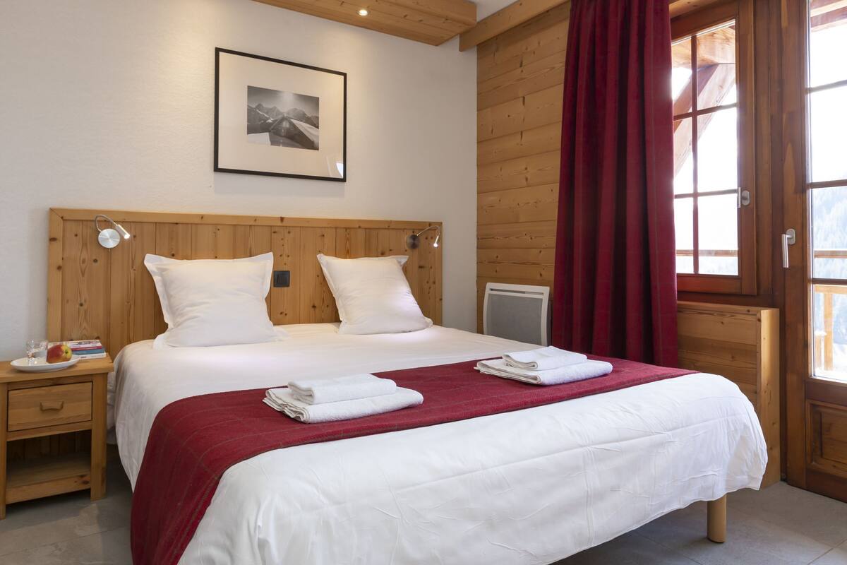 A double bed with white bedding, a red throw blanket, and two pillows. There is a framed picture on the wall above the bed, a wooden headboard, and a window with red curtains.