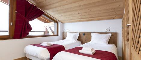 Habitación con dos camas, techo de madera y ventana con cortinas rojas.