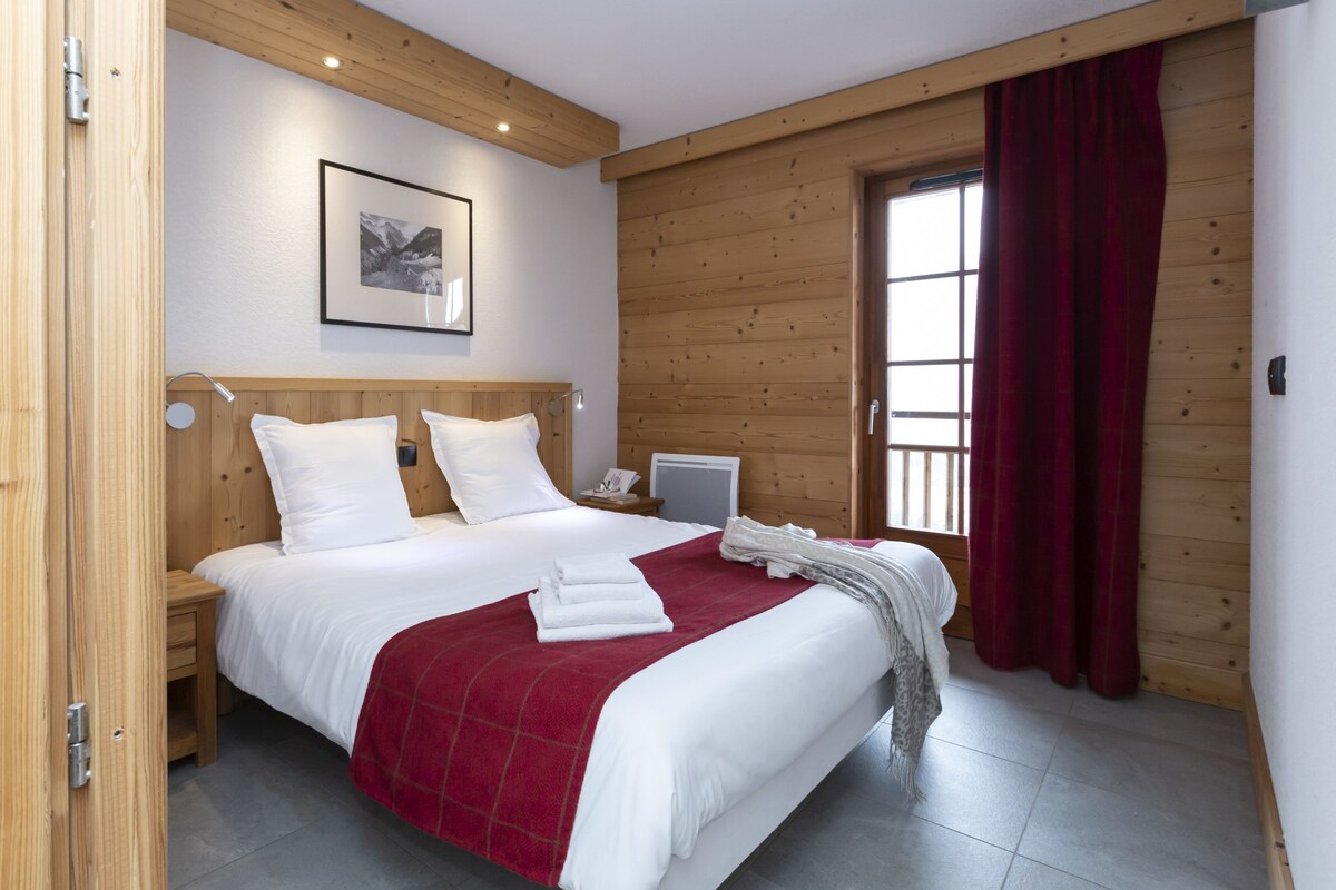 A bedroom with a wooden headboard, a bed with white bedding and a red blanket, a bedside table with a lamp, and a window with red curtains.