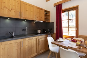 A kitchen with wooden cabinets, a black countertop, a window with a red curtain, and a breakfast table with breakfast items. - Residence Odalys Les Fermes De Chatel (Chatel)
