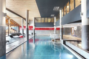 A modern indoor swimming pool with clear water, lounge chairs, and a high ceiling with wooden beams.