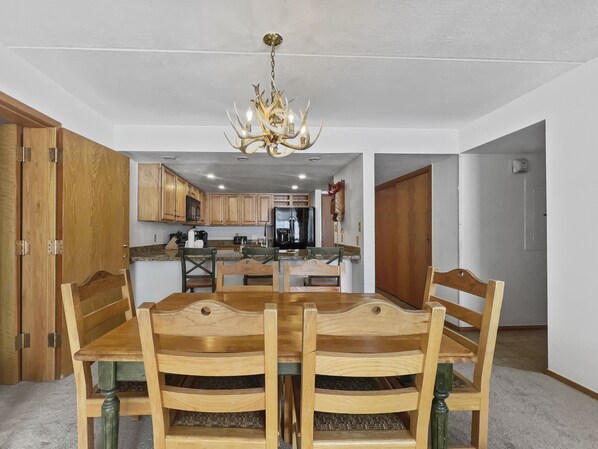 A dining area with a wooden table and chairs, a chandelier, and a kitchen in the background. - Ski in/ Ski Out Condo, Perfect Location, Family Friendly, Great View (Copper Mountain)