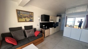 En moderne stue med sofa, et lille hvidt bord, et tekøkken med skabe og et fjernsyn, samt en balkon med udsigt.