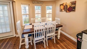 Une salle à manger avec une table en bois et des chaises blanches, de grandes fenêtres et une œuvre d’art accrochée au mur.