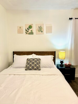 A neatly made bed with white linens and a patterned pillow, a bedside table with a lamp and a basket, and three wall art pieces above the bed. - Cozy 3-Bedroom Home w/ Covered Deck. Pet-Friendly! (Lawrenceville)
