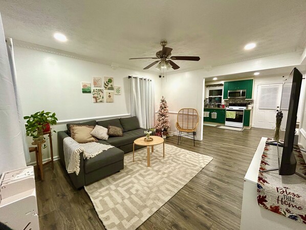 A living room with a sofa, a small table, and a Christmas tree. There is a kitchen in the background with green cabinets and a microwave. - Cozy 3-Bedroom Home w/ Covered Deck. Pet-Friendly! (Lawrenceville)