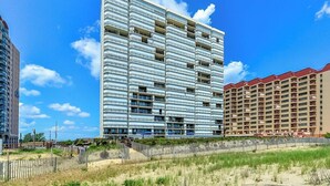 A modern high-rise hotel with multiple balconies, a sandy beachfront, and a clear blue sky.
