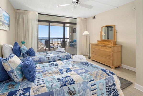 A hotel room with two beds, a wooden dresser, a ceiling fan, a large window with a view of the beach, and a balcony with chairs.