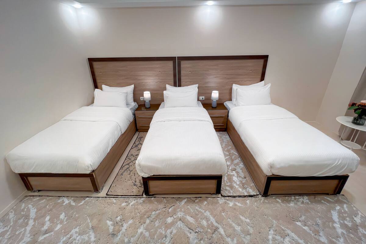 A hotel room with three single beds, a wooden headboard, bedside tables with lamps, and a small round table with a vase of flowers.