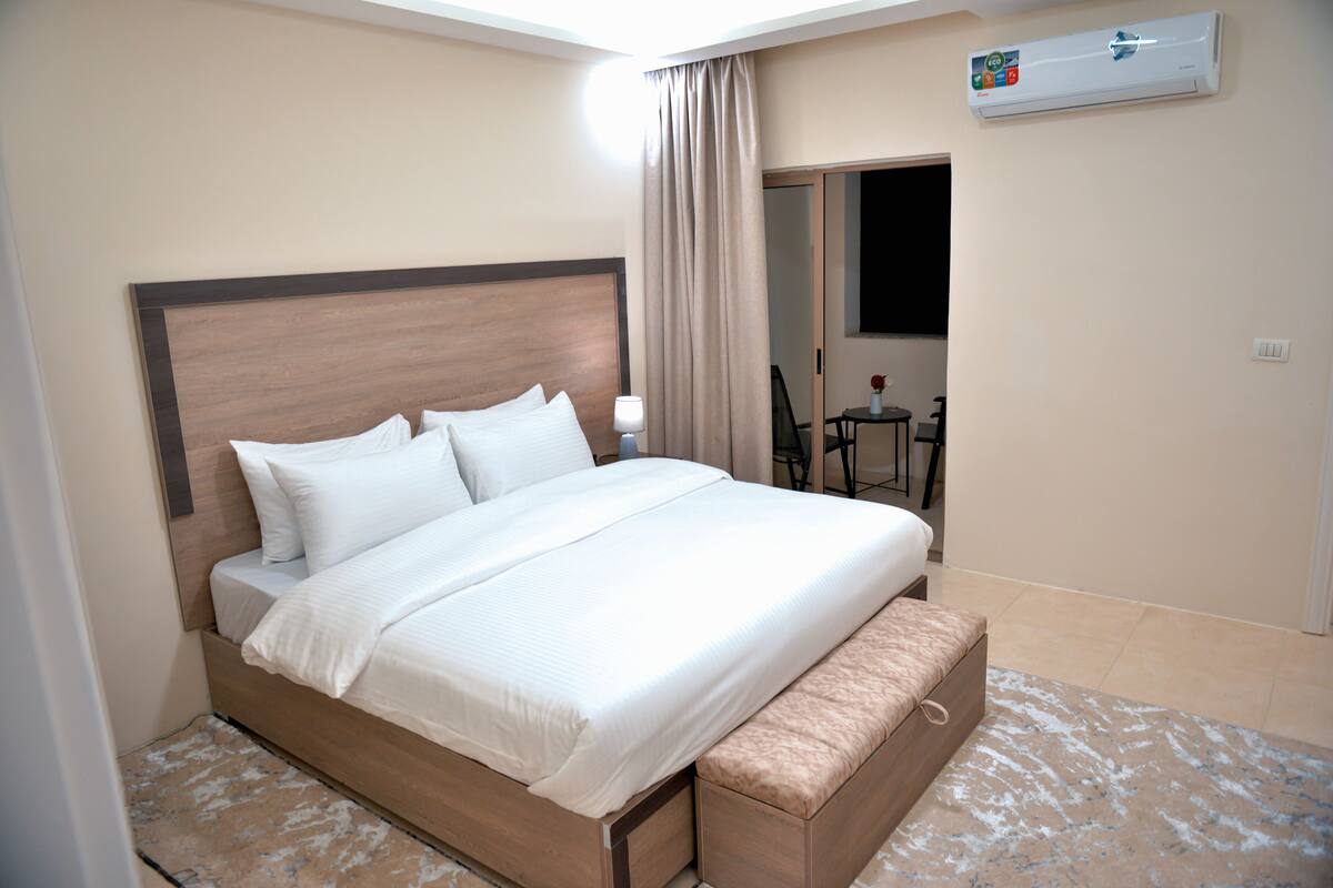A hotel room with a large bed, a bedside table, a chair, a small table, and an air conditioning unit.