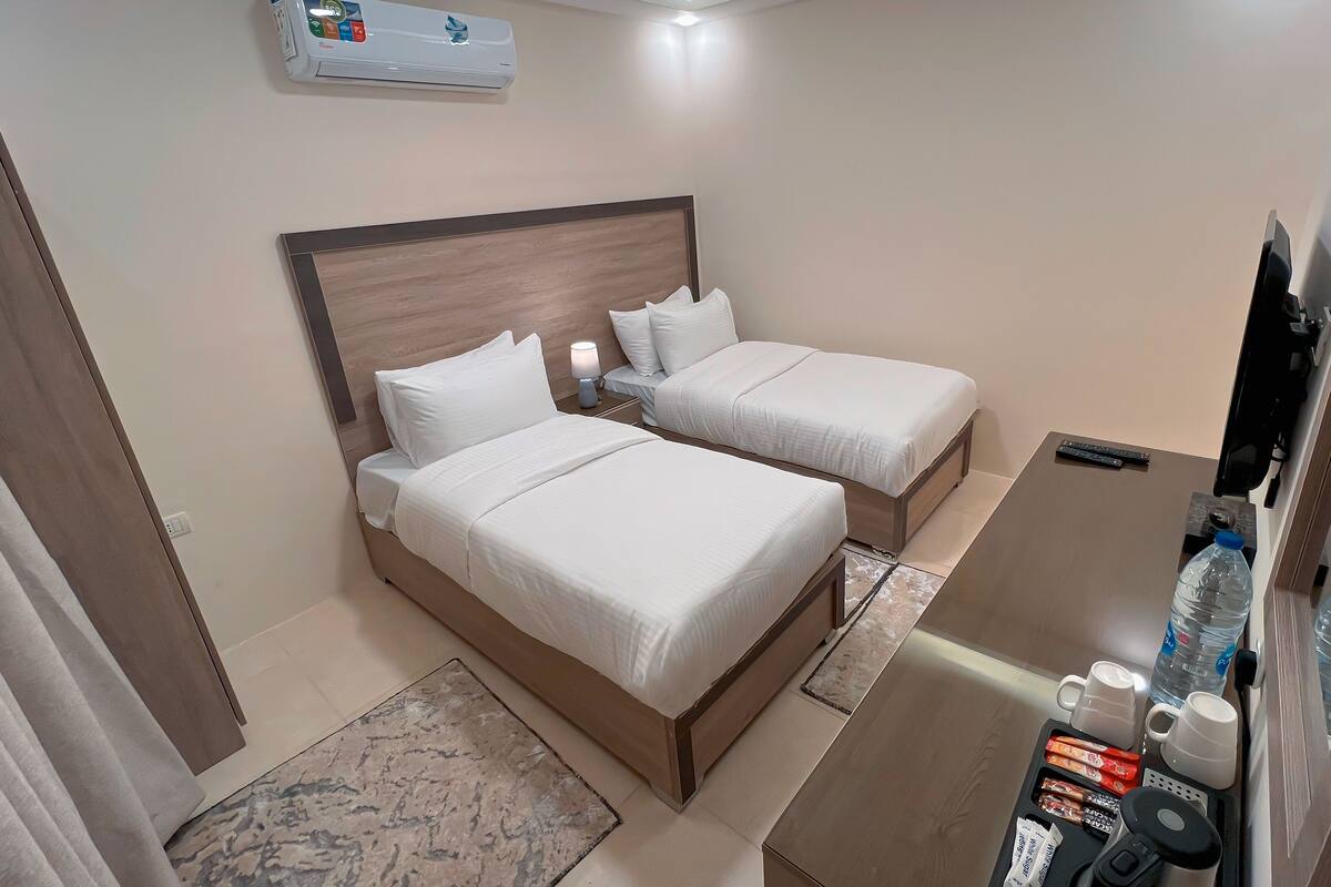 A hotel room with two beds, a desk with a water bottle, cups, and a remote control.