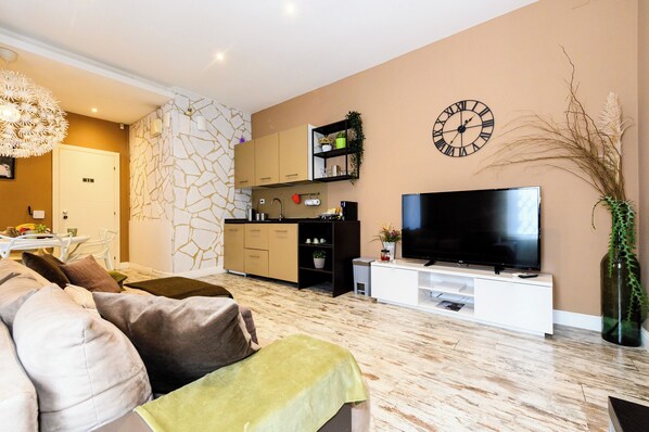 A modern living room with a sofa, a TV, and a kitchen area with cabinets and a clock. - A-Rome Suite (Rome)