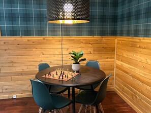 A corner room with a wooden wall, a round table with chairs, a chessboard, and a hanging light fixture.