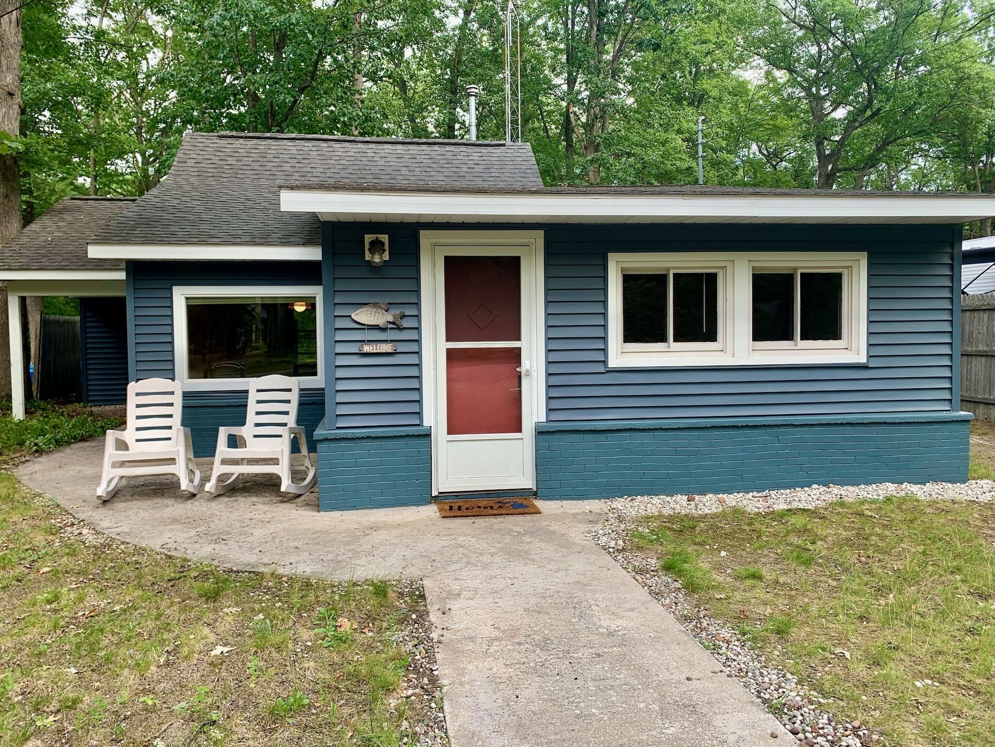 A single-story house with a dark blue exterior and a white door. There are two white plastic chairs outside, and a welcome sign with a fish decoration.