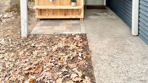 A wooden outdoor seating area with a bench and a towel rack, a small potted plant, and a concrete pathway. - Cabin @ Torch with Lake Access, Kayaks & Outdoor Shower! (Rapid City)