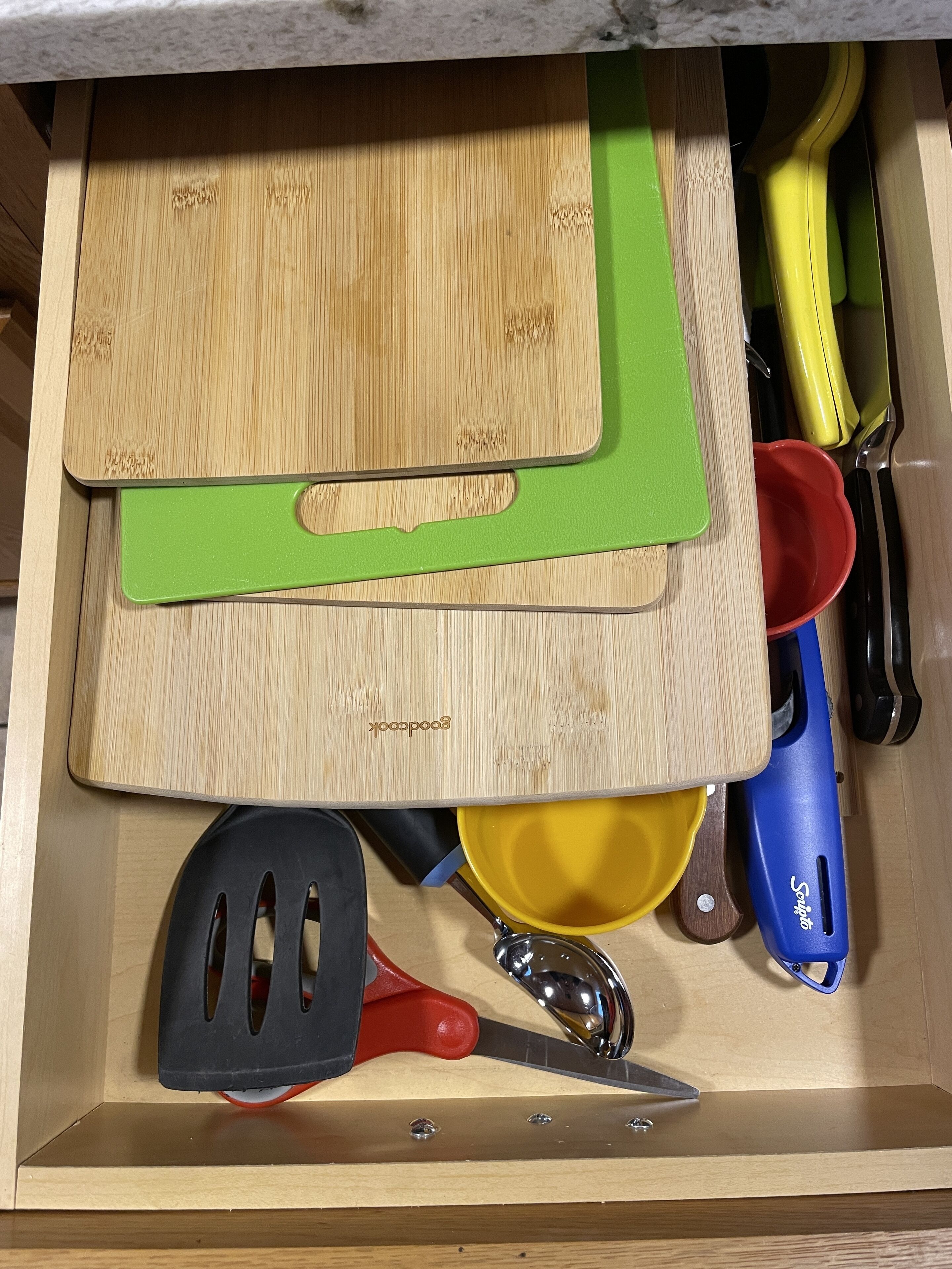 Un cajón con diversos utensilios y herramientas de cocina, incluyendo una tabla de cortar de madera, un tapete de corte verde, una taza medidora amarilla, una espátula azul y una espátula roja.