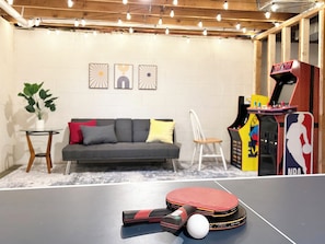 A ping pong table with paddles and a ball, a sofa with cushions, a small table with a plant, and a retro arcade machine.