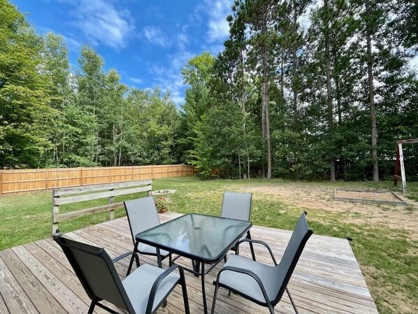 An outdoor patio with a glass table and four chairs, surrounded by a wooden deck and a wooden fence.