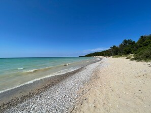 En strand med småsten, klart turkosfärgat vatten och ett skogsområde i bakgrunden.