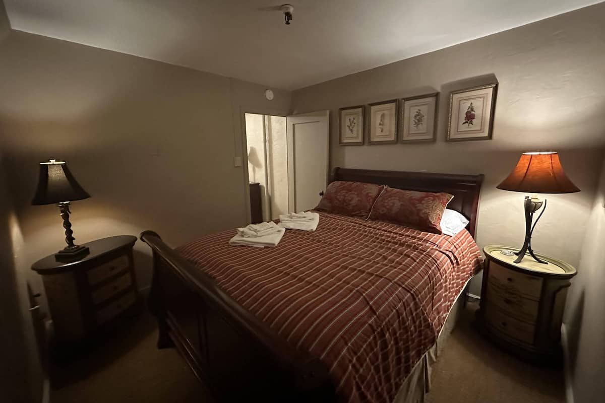 Un lit bien fait, recouvert d’une couvre-lit à rayures, deux tables de chevet avec des lampes, une petite table en bois et des tableaux encadrés accrochés au mur.