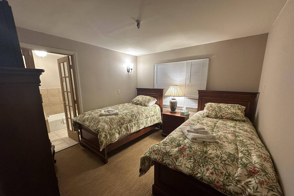 Une chambre avec deux lits, une table de chevet, une lampe et une fenêtre avec des stores.