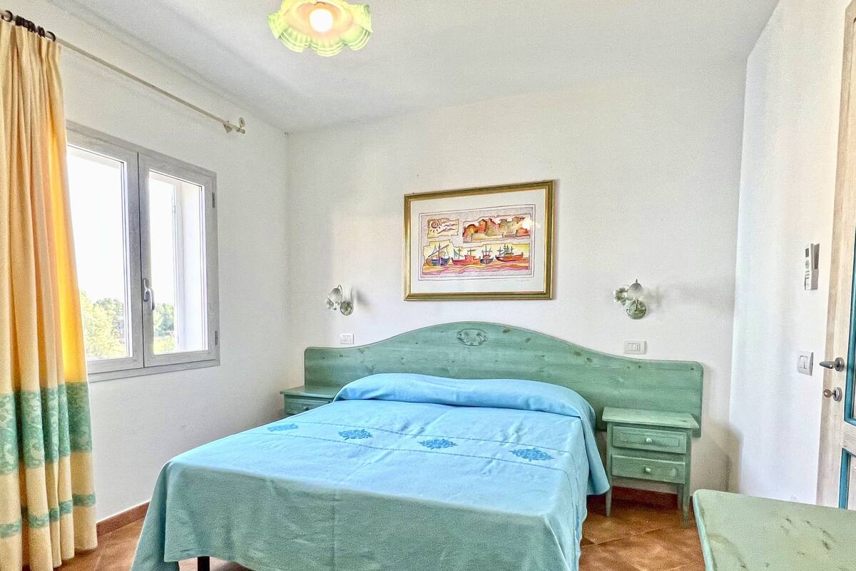 Une chambre à coucher comprenant un lit, une table de chevet, une commode, une fenêtre avec des rideaux, un luminaire fixé au mur et un tableau encadré.