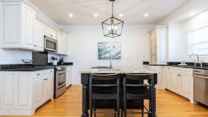 A modern kitchen with white cabinetry, a central island with bar stools, a stainless steel range hood, and a pendant light. - Sunset Soiree-501 A 22nd St · Sunset Soiree in Virginia Beach (Virginia Beach)