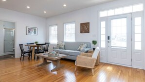 A spacious living room with a grey sofa, wooden floor, and a dining area with a table and chairs. - Sunset Soiree-501 A 22nd St · Sunset Soiree in Virginia Beach (Virginia Beach)