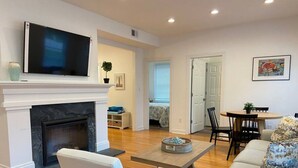 A living room with a fireplace, a flat-screen TV mounted above it, a dining area with a table and chairs, and a bedroom visible through an open door. - Sunset Soiree-501 A 22nd St · Sunset Soiree in Virginia Beach (Virginia Beach)