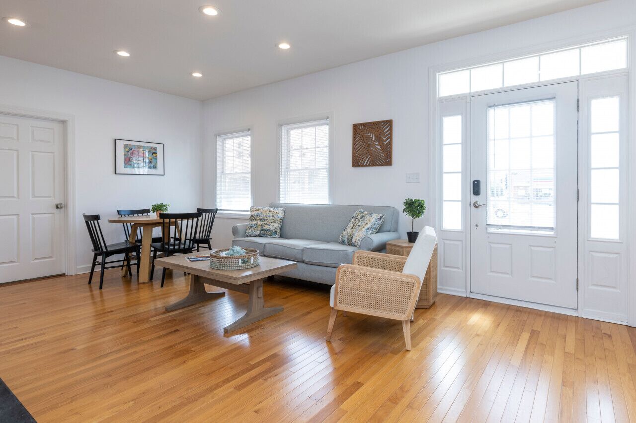 A spacious living room with a grey sofa, a dining table with chairs, and a large window.