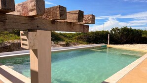 A clear pool with a wooden pergola overhead, surrounded by a sandy area and greenery.