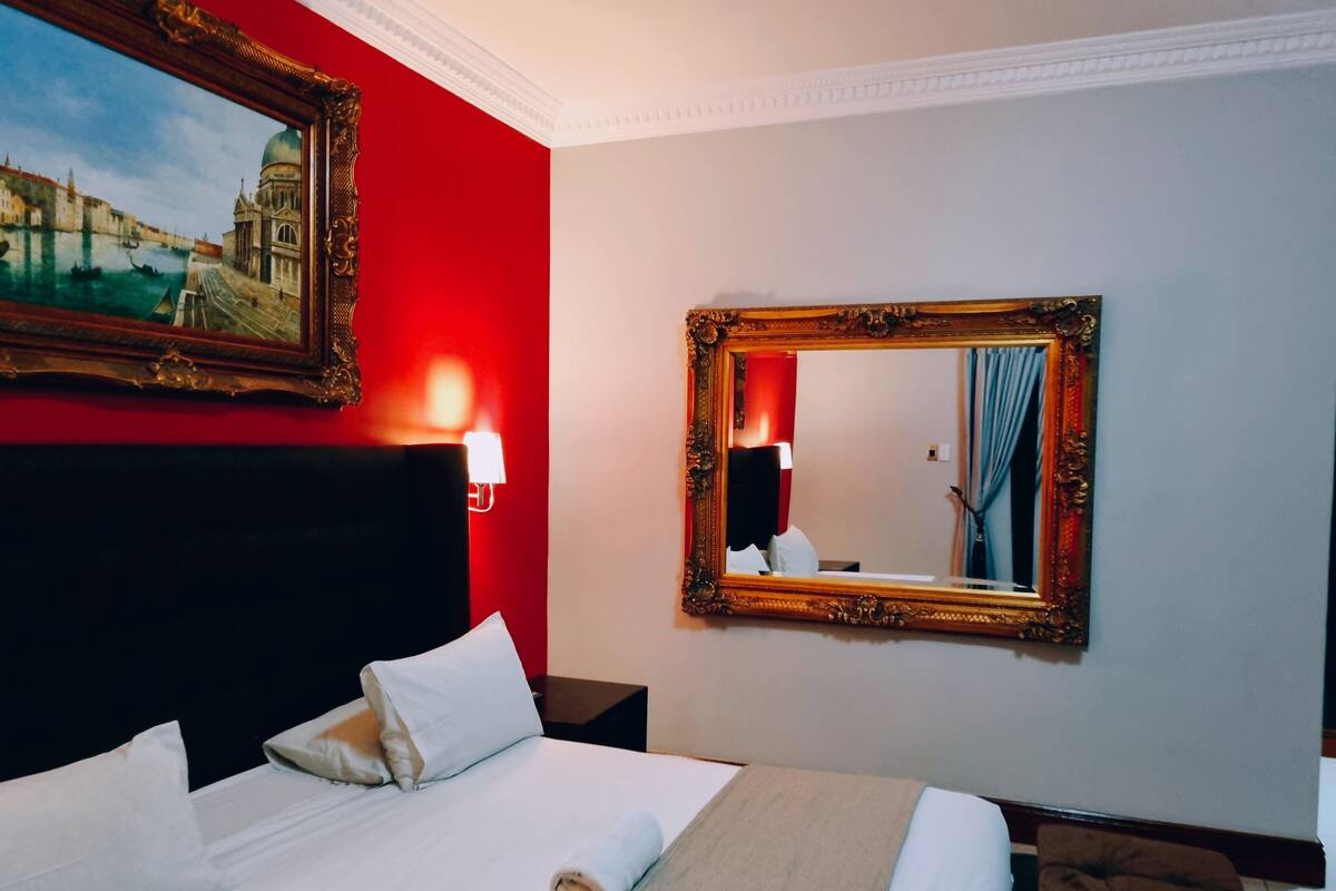 Una habitación de hotel con una cama grande, cabecera, espejo fijado a la pared y un cuadro de un paisaje urbano.