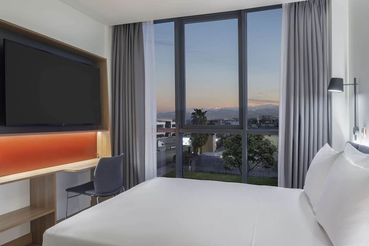 Een hotelkamer met een bed, een bureau met een stoel, een aan de muur bevestigde televisie en een groot raam met uitzicht op de stad en de bergen.