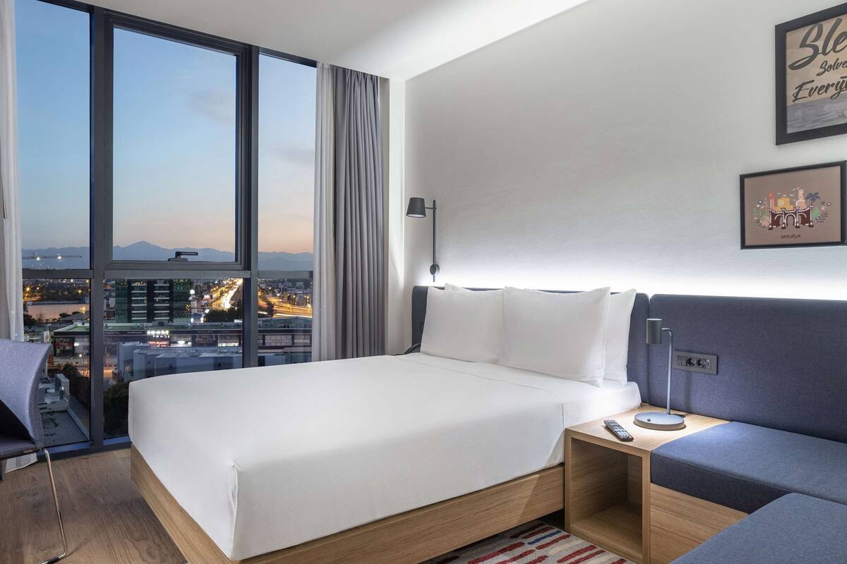 Een moderne hotelkamer met een groot bed, een bureau en een bank. Er zijn grote ramen met uitzicht op de stad.