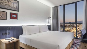 Hotelkamer met een groot bed, een bureau, een stoel en uitzicht op een stadslandschap door grote ramen.