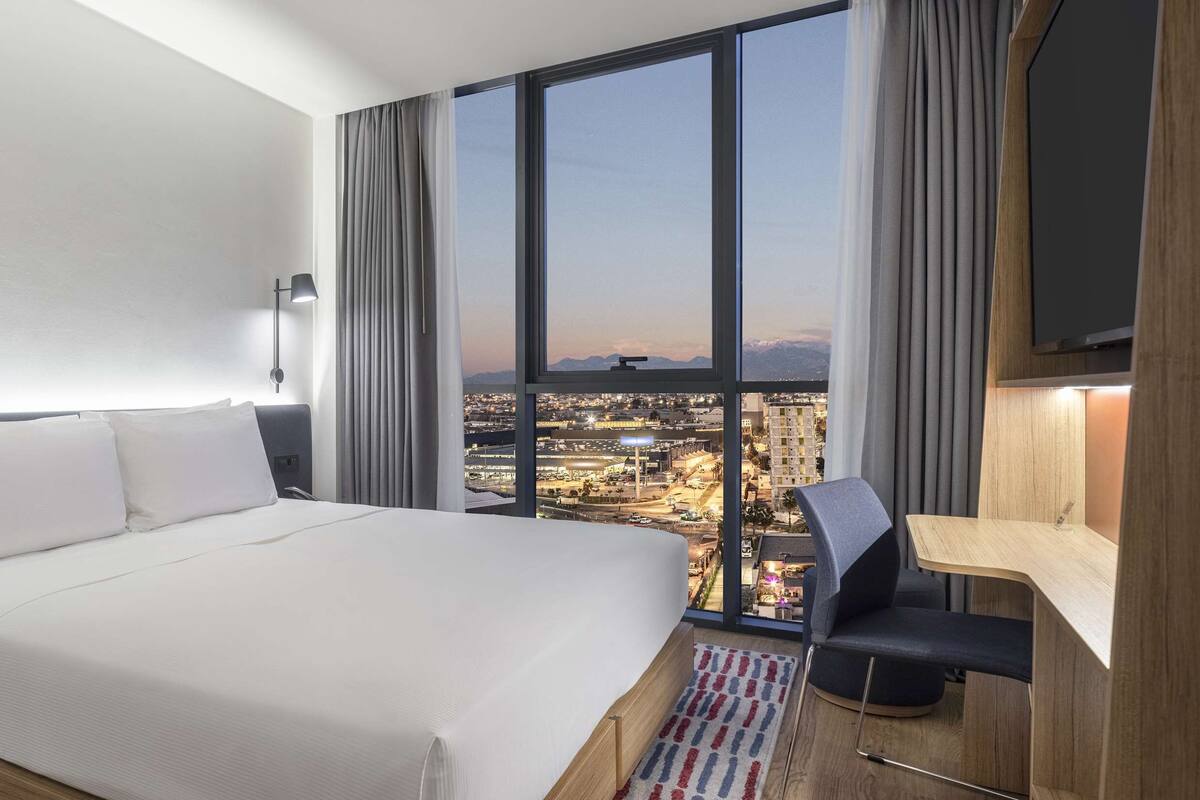 Hotelkamer met een groot bed, een bureau, een stoel en uitzicht op een stadslandschap door grote ramen.
