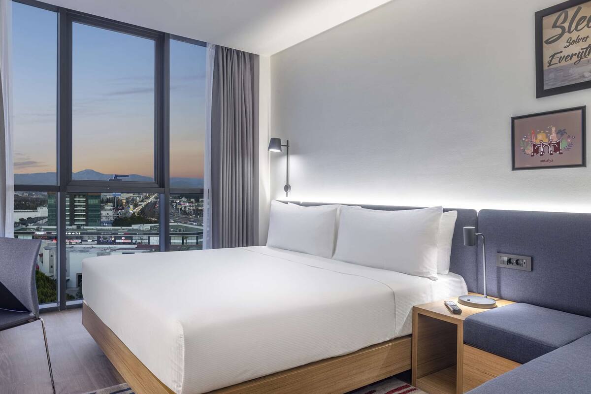 Een moderne hotelkamer met een groot bed, een bureau en een bank. Er zijn grote ramen met uitzicht op de stad.
