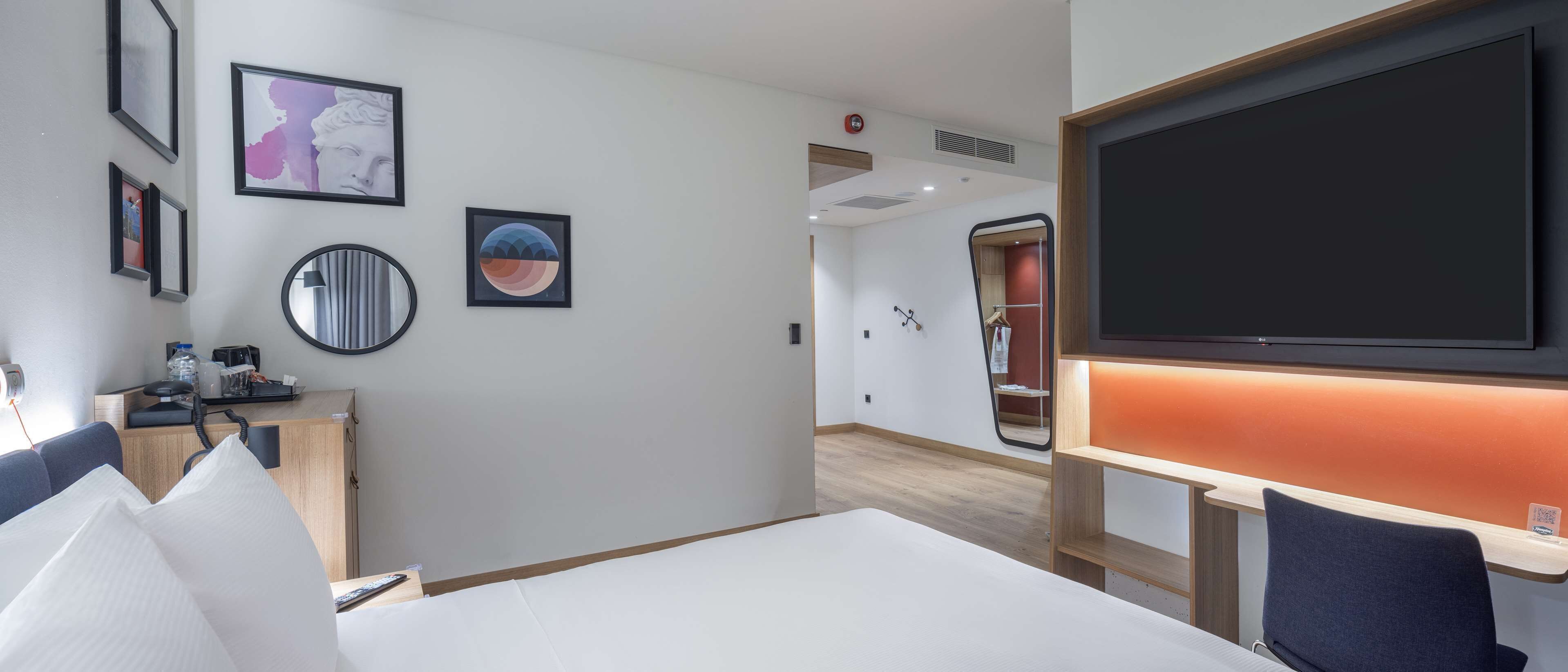 Une chambre d’hôtel avec un lit, un bureau avec un ordinateur, une télévision et un miroir. Les murs sont ornés de tableaux encadrés et il y a un meuble en bois.