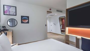 A hotel room with a bed, a desk with a computer, a TV, and a mirror. There are framed pictures on the wall and a wooden cabinet.