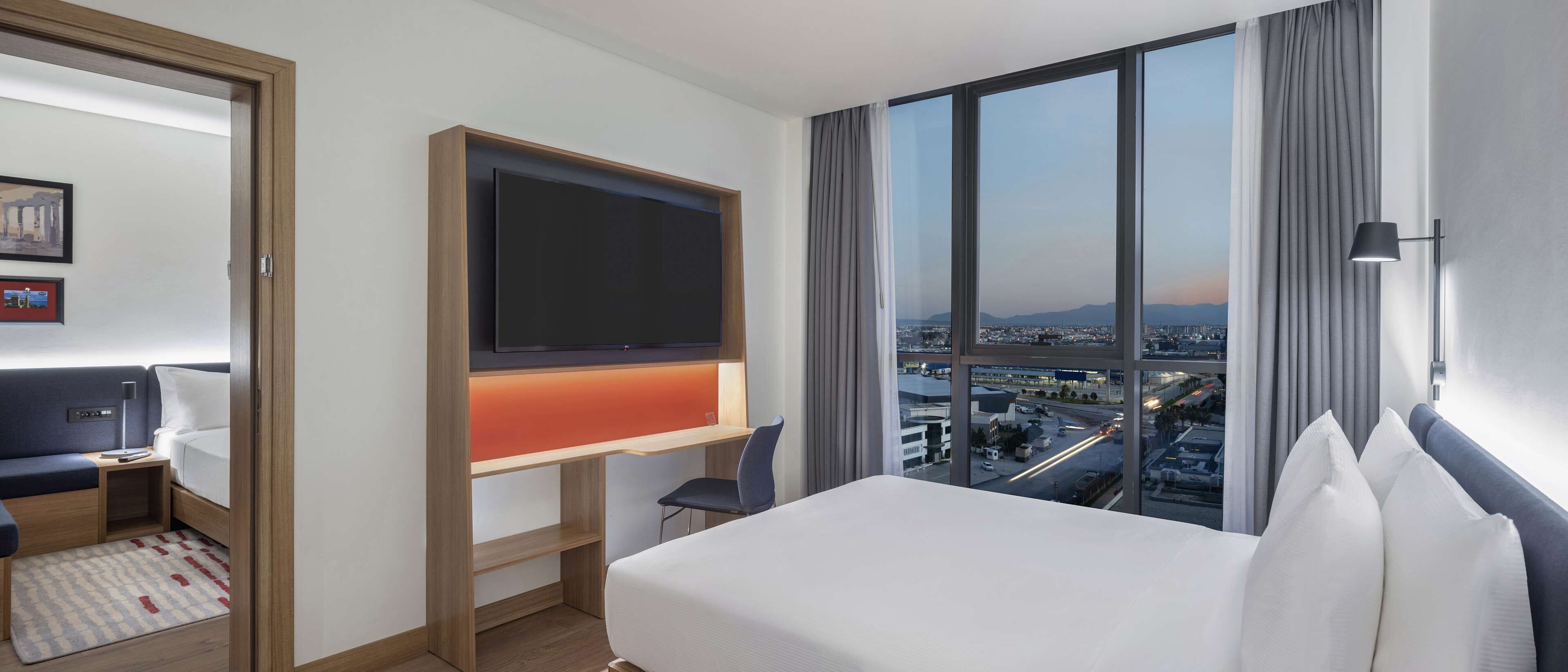 Habitación de hotel con cama, escritorio, televisor montado en la pared y un amplio ventanal con vistas a la ciudad.