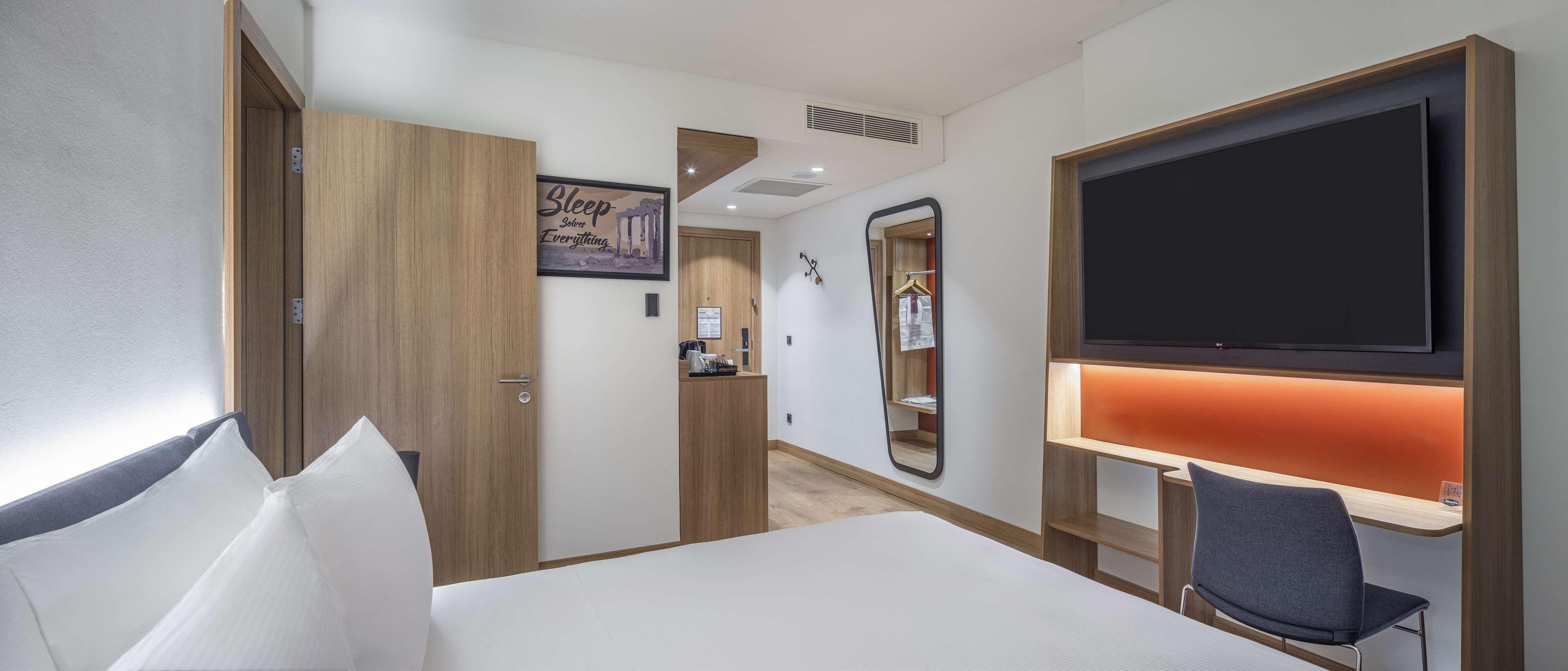 A hotel room with a bed, a desk with a chair, a TV, a mirror, and a wardrobe.