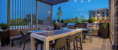 An outdoor patio with a long table and chairs, a barbecue grill, and string lights.