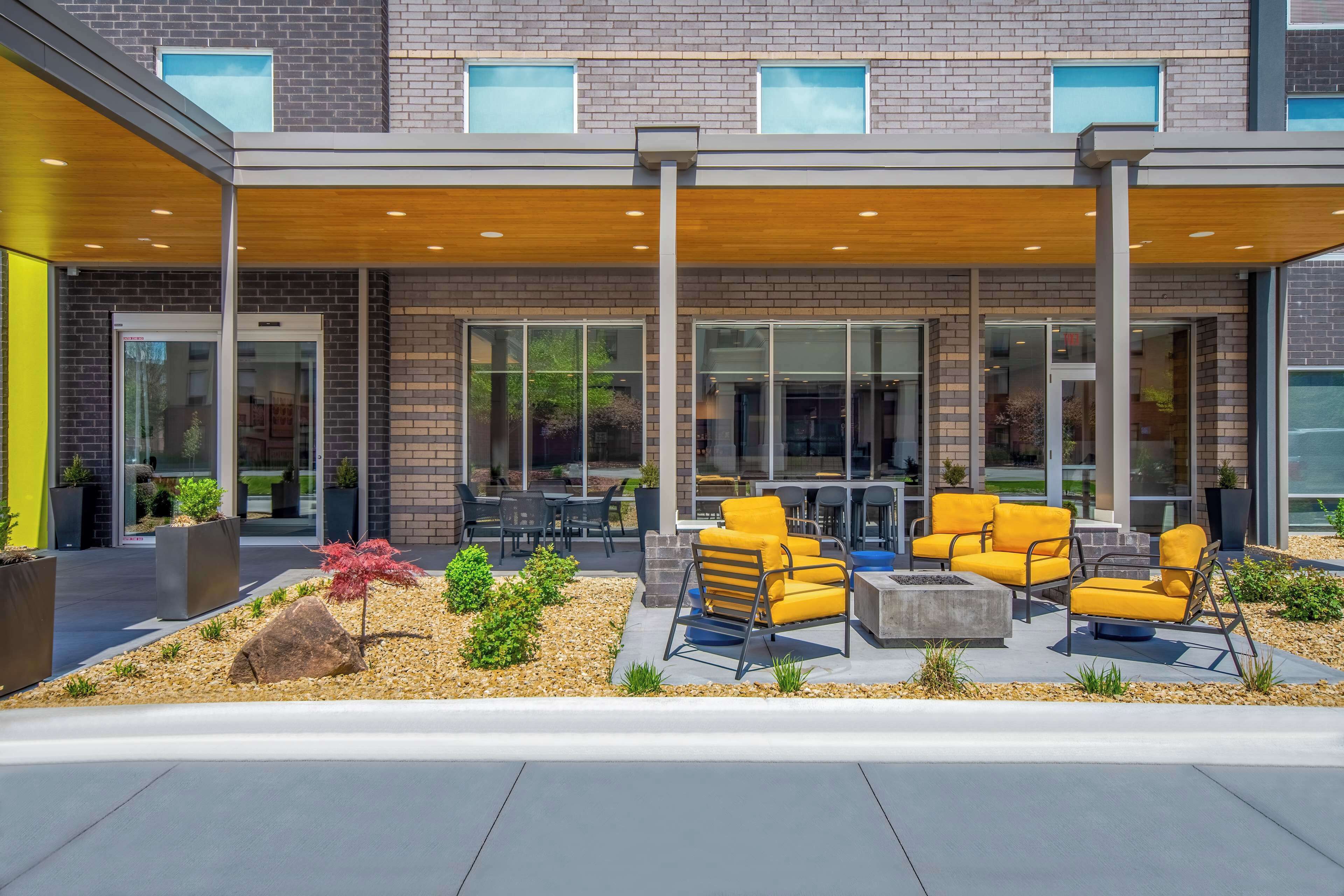 An outdoor seating area with yellow chairs and tables, a brick building with large windows, and a covered patio area.