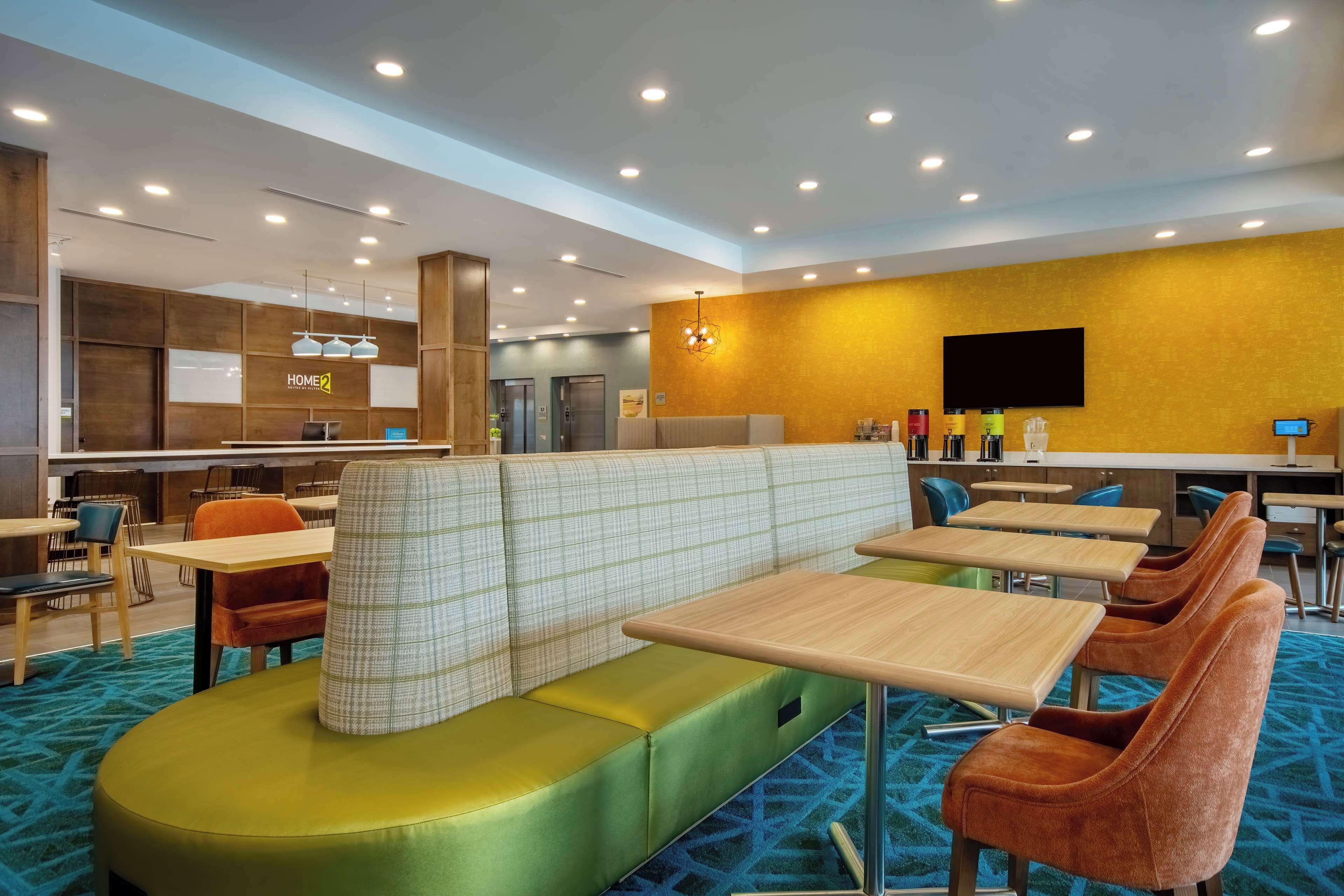 A modern hotel lobby with seating areas, a reception desk, and a kitchen in the background.