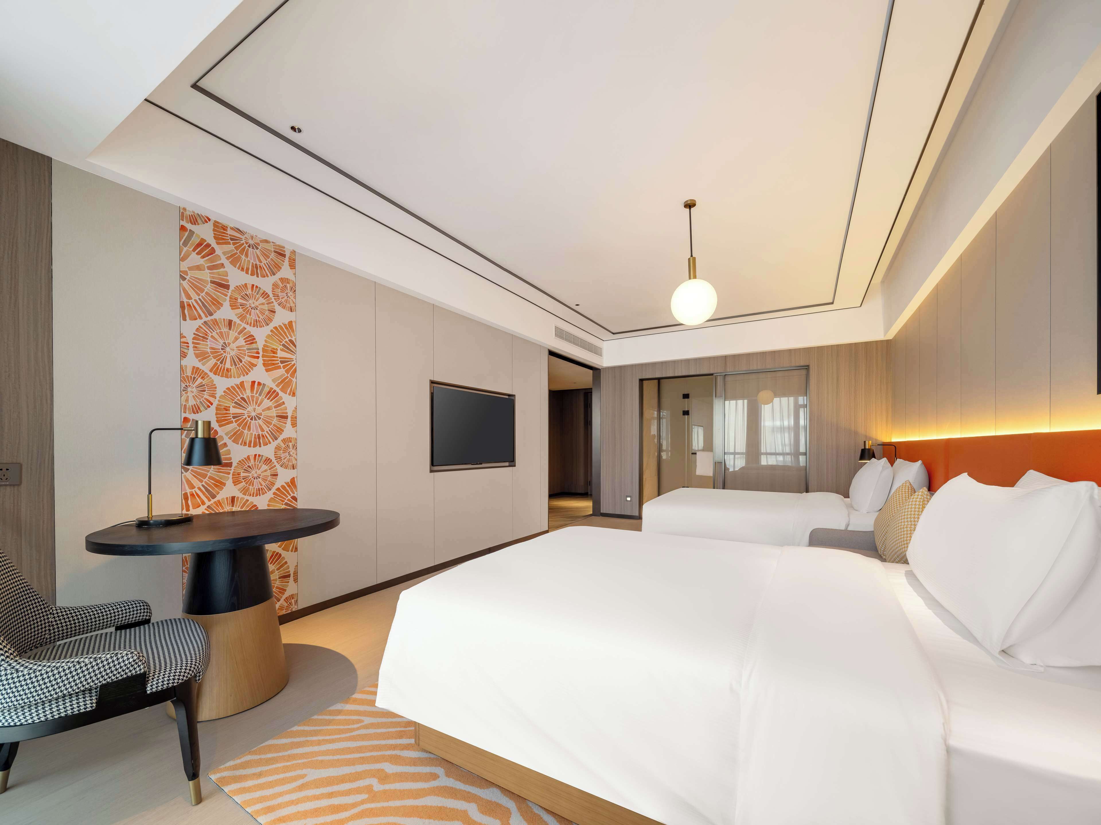 A modern hotel room with a large bed, a desk with a chair, a TV, and a unique orange patterned wall.