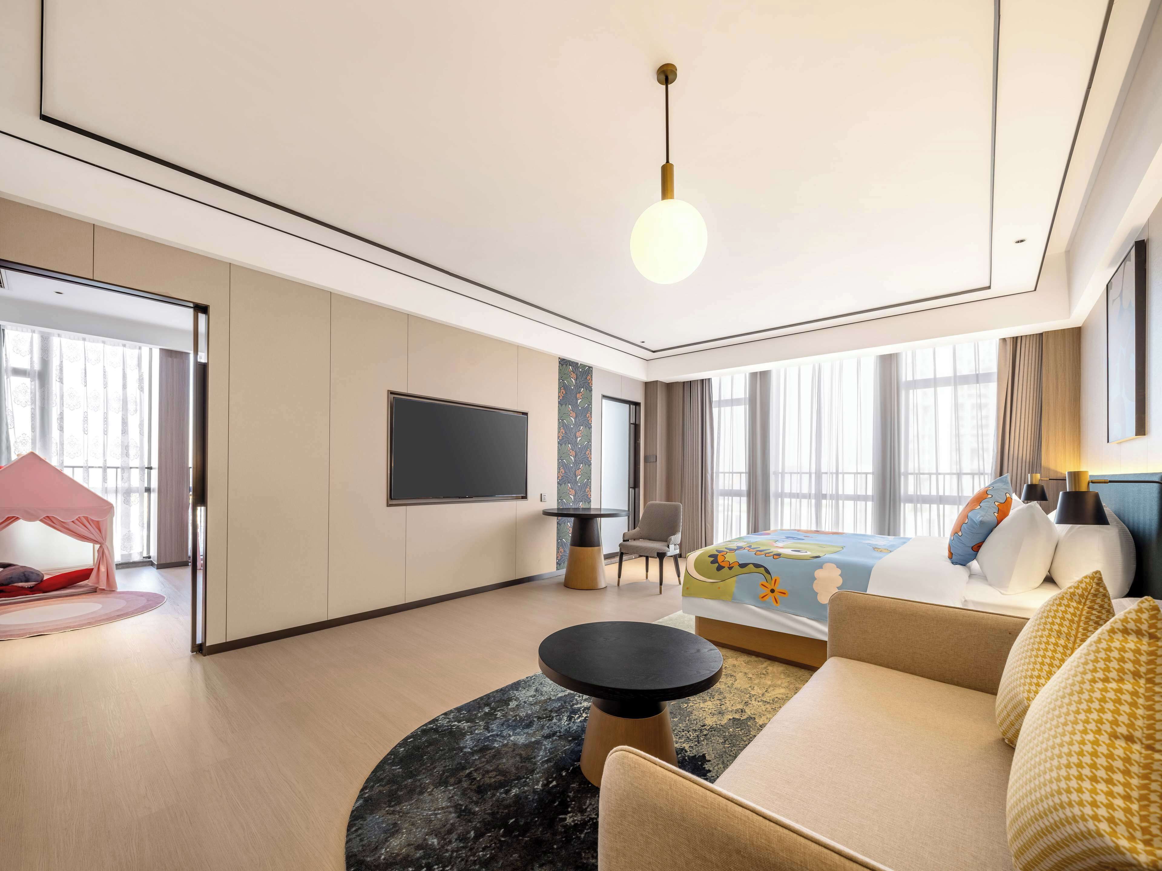 A modern hotel room with a large bed, a seating area with a sofa and armchairs, a flat-screen TV mounted on the wall, and a large window offering a view.
